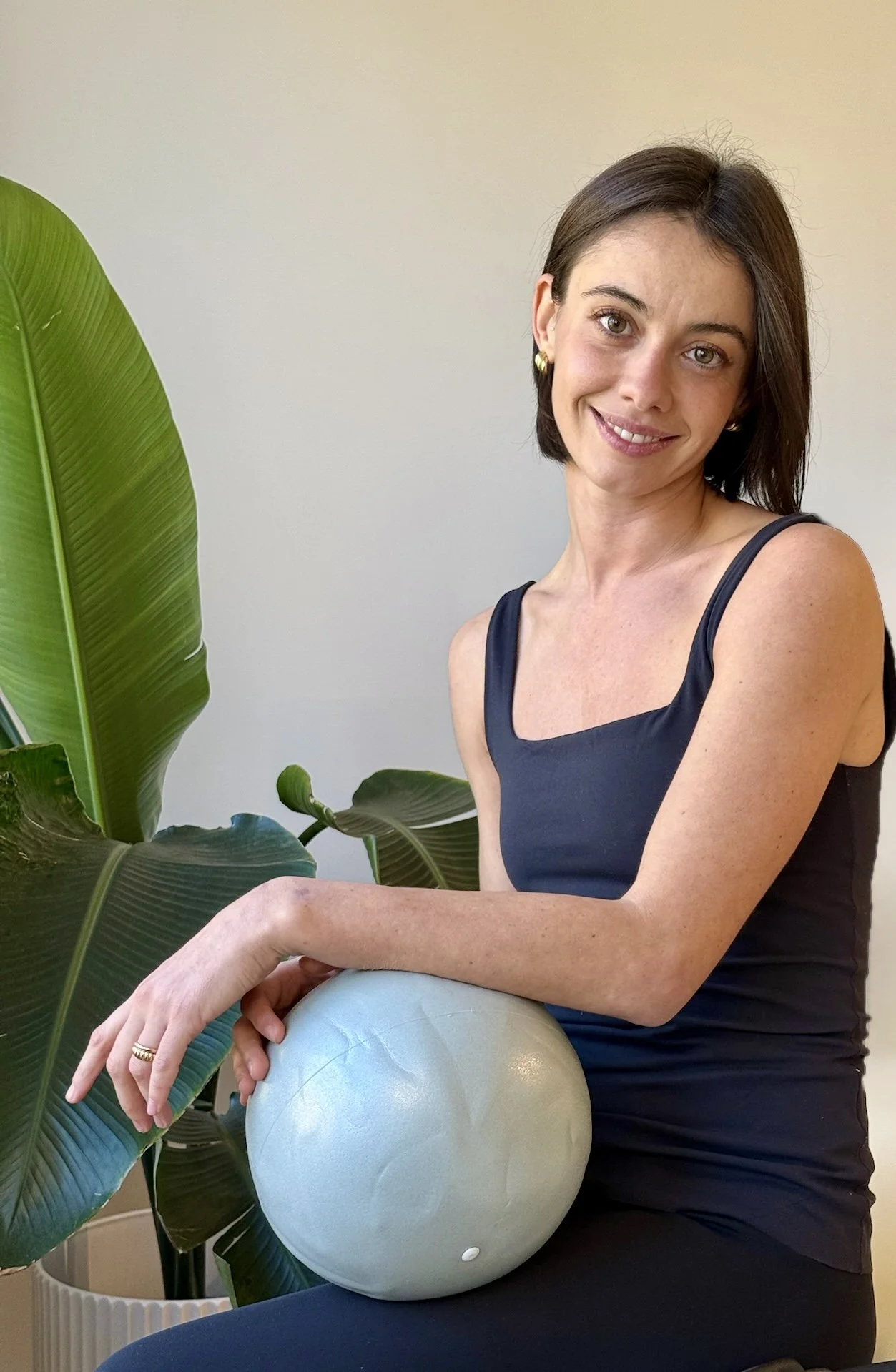 A woman with short brown hair, wearing a black sleeveless top, sitting and holding a light gray exercise ball, with large green indoor plants nearby.