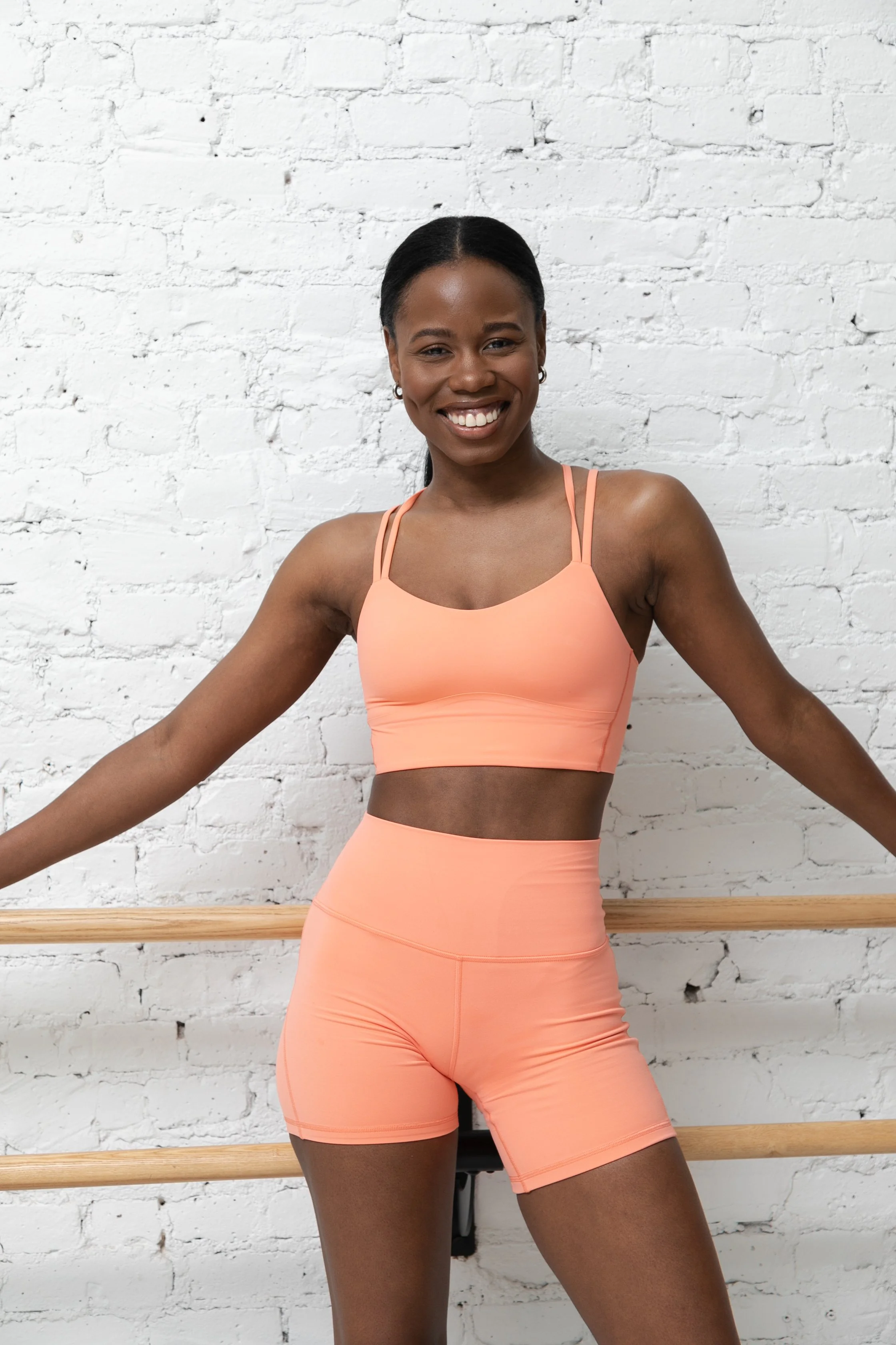 Smiling woman in peach workout outfit standing in front of a white brick wall with ballet bar