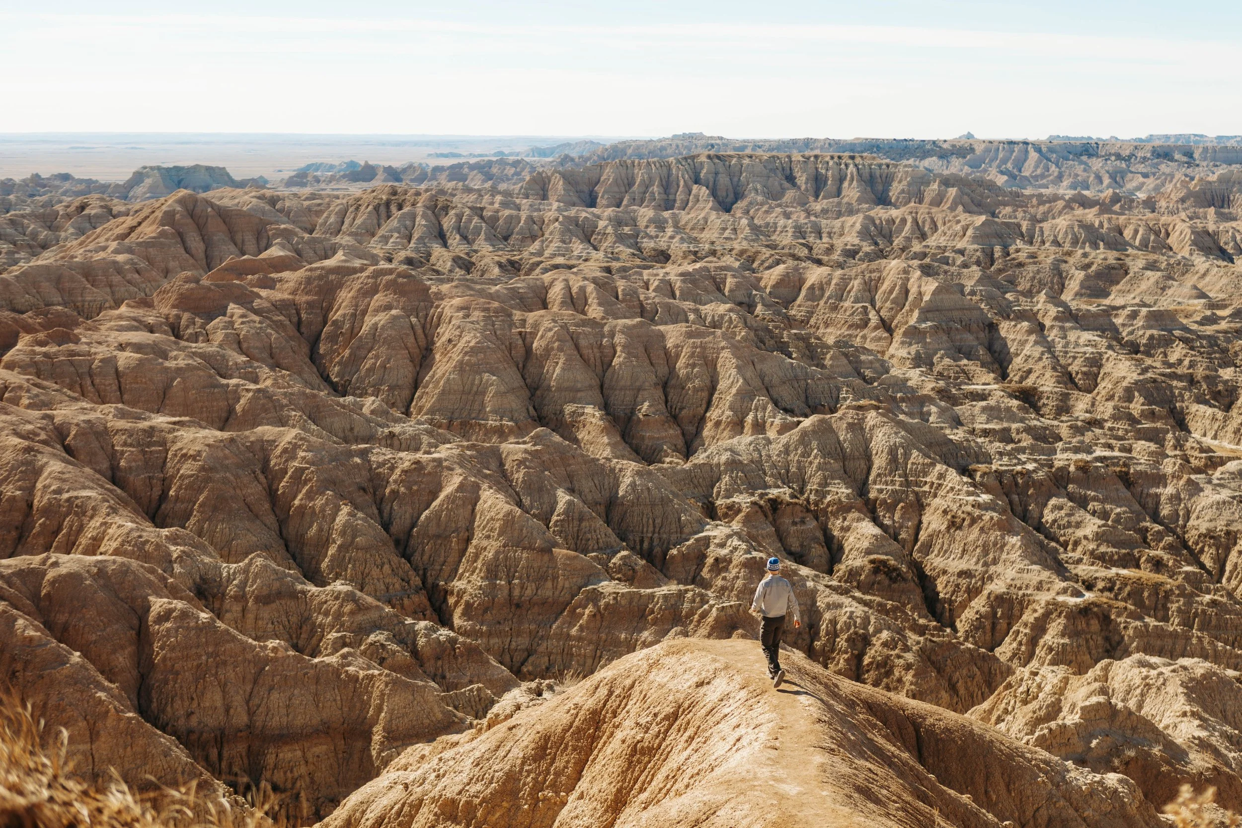 A person walking along a narrow trail on a rugged, eroded canyon landscape with layered rock formations and a vast, open sky in the background in Badlands National Park.
