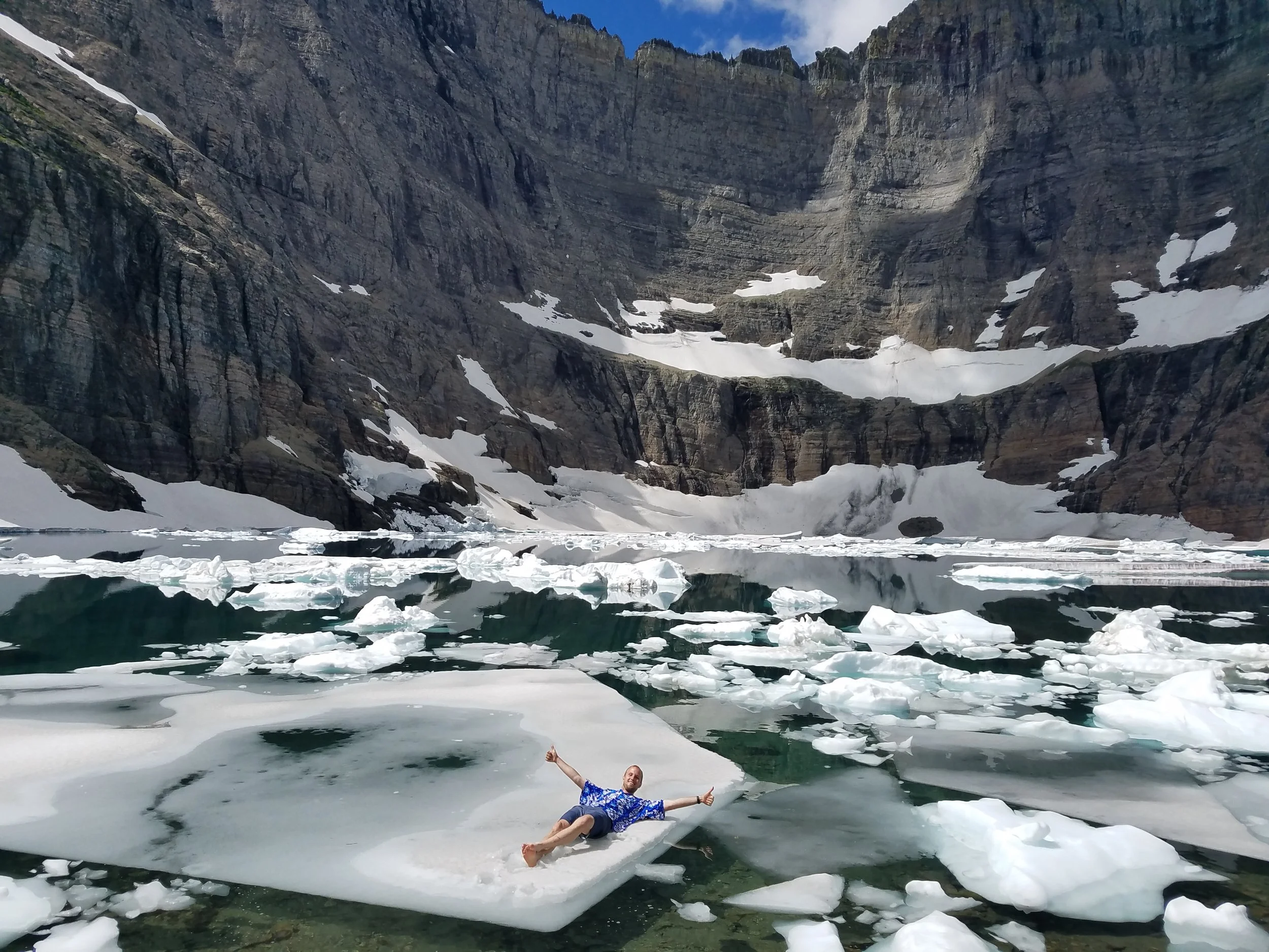 A person in a blue Hawaiian shirt and shorts lying on floating iceberg with steep rocky mountains and patches of snow behind at Iceberg Lake in Glacier National Park, Montana.