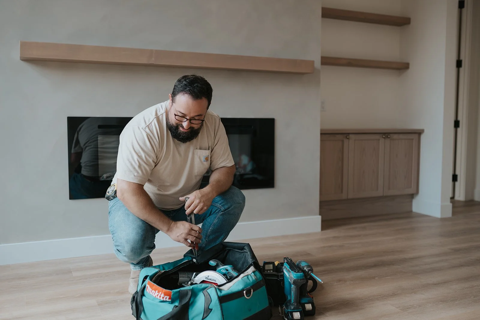 A man with glasses and a beard, wearing a beige t-shirt and jeans, is kneeling and packing a teal Makita tool bag with various power tools and accessories.