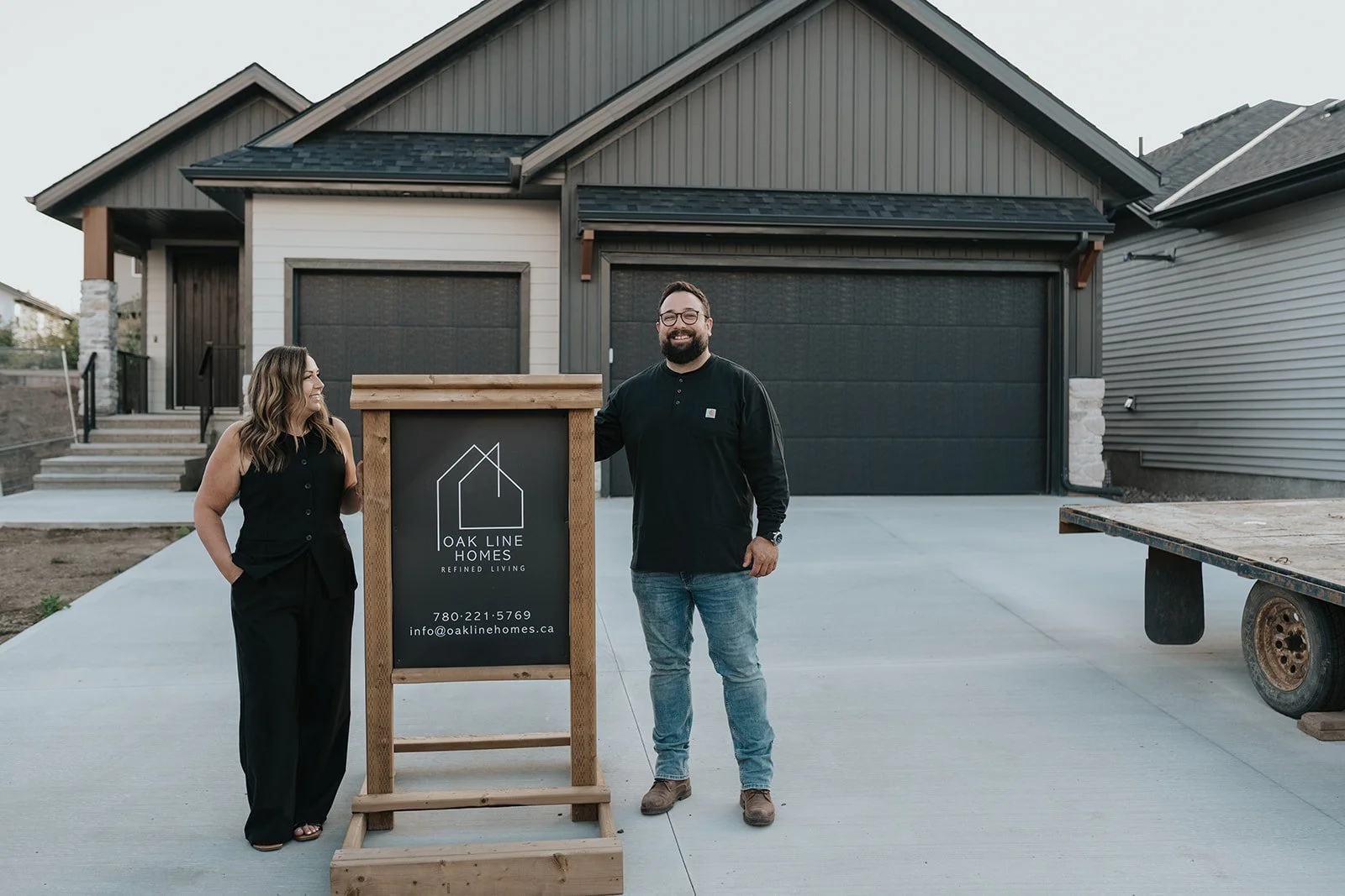 Two people standing in front of a new house, smiling and looking at each other, with a sign for Oak Line Homes in between them.