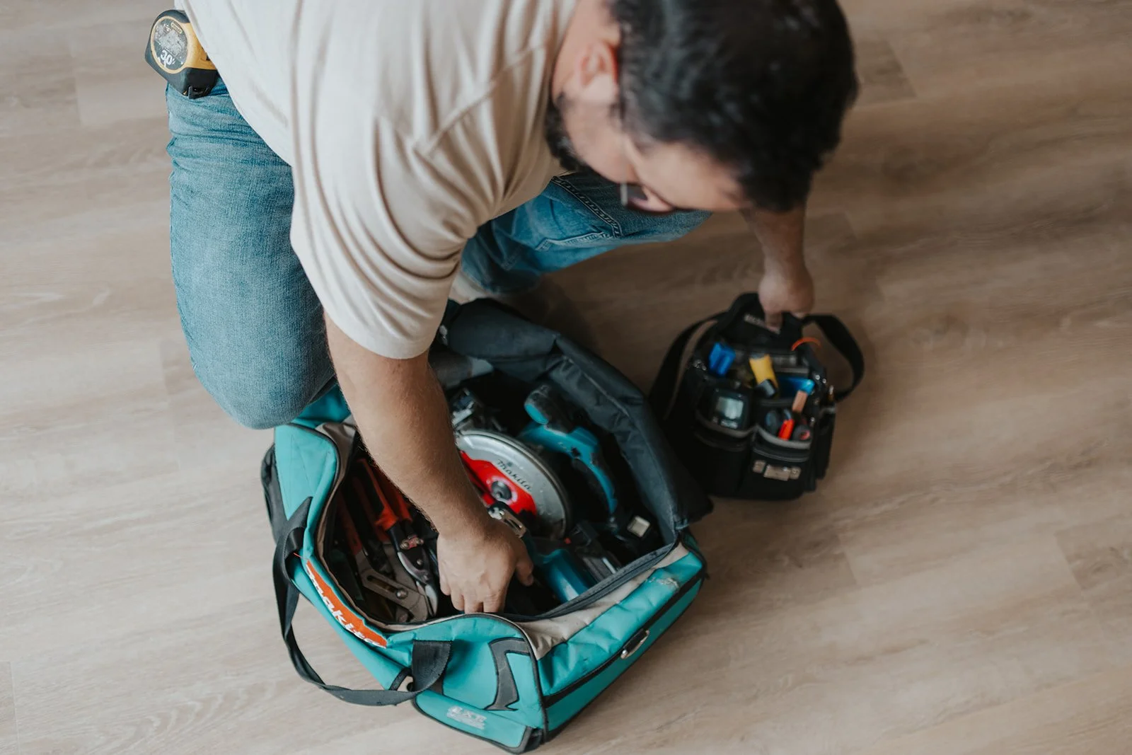 A man is kneeling on a wooden floor, packing tools into a blue and black tool bag. The tools include a power saw, wrenches, and screwdrivers. There is another black tool bag nearby with various tools inside.