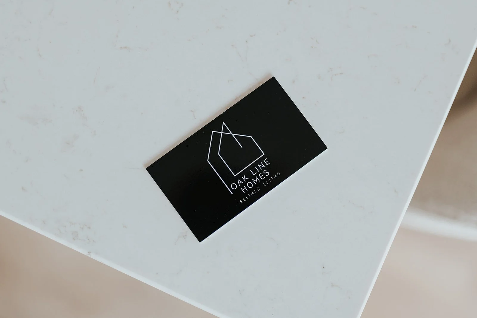 Black business card with white geometric house outline, displaying 'OAKLINE HOMES' and 'REFINED LIVING' on a white surface.