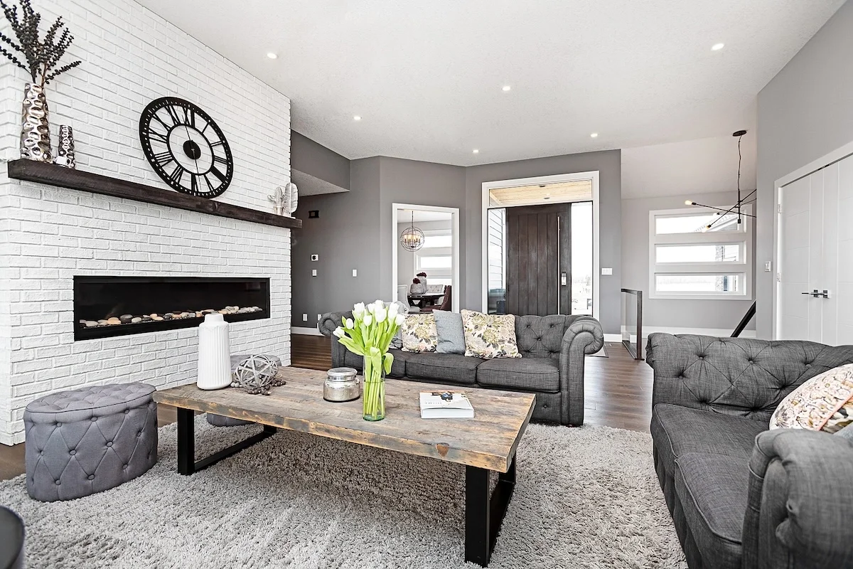 A modern living room with gray walls, a white brick fireplace, and a dark wooden door. The room has gray tufted sofas, a rustic wooden coffee table with a vase of white tulips, and decorative pillows. There's a large black wall clock and a bookshelf on the white brick wall. The space is illuminated by recessed ceiling lights and natural light through the windows.