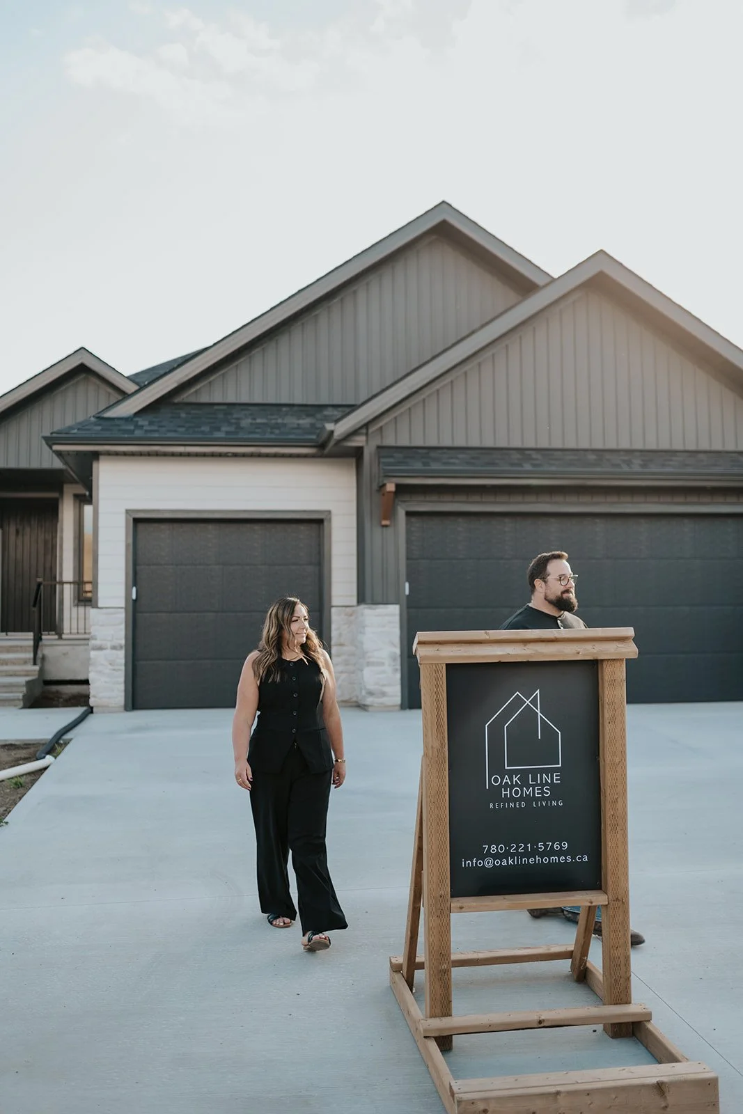 Two people walking outside in front of a modern house with a black garage door. There is a wooden signboard with the logo and contact information for Oak Line Homes, a homebuilding company.