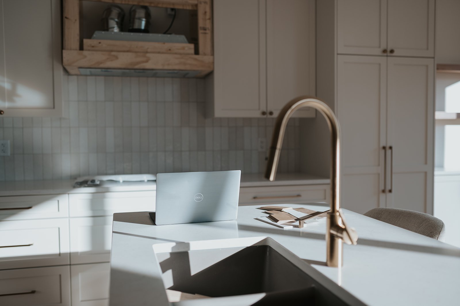 A modern kitchen with white cabinets, a light-colored tile backsplash, a gold kitchen faucet, a laptop on the white kitchen island, and some papers nearby.