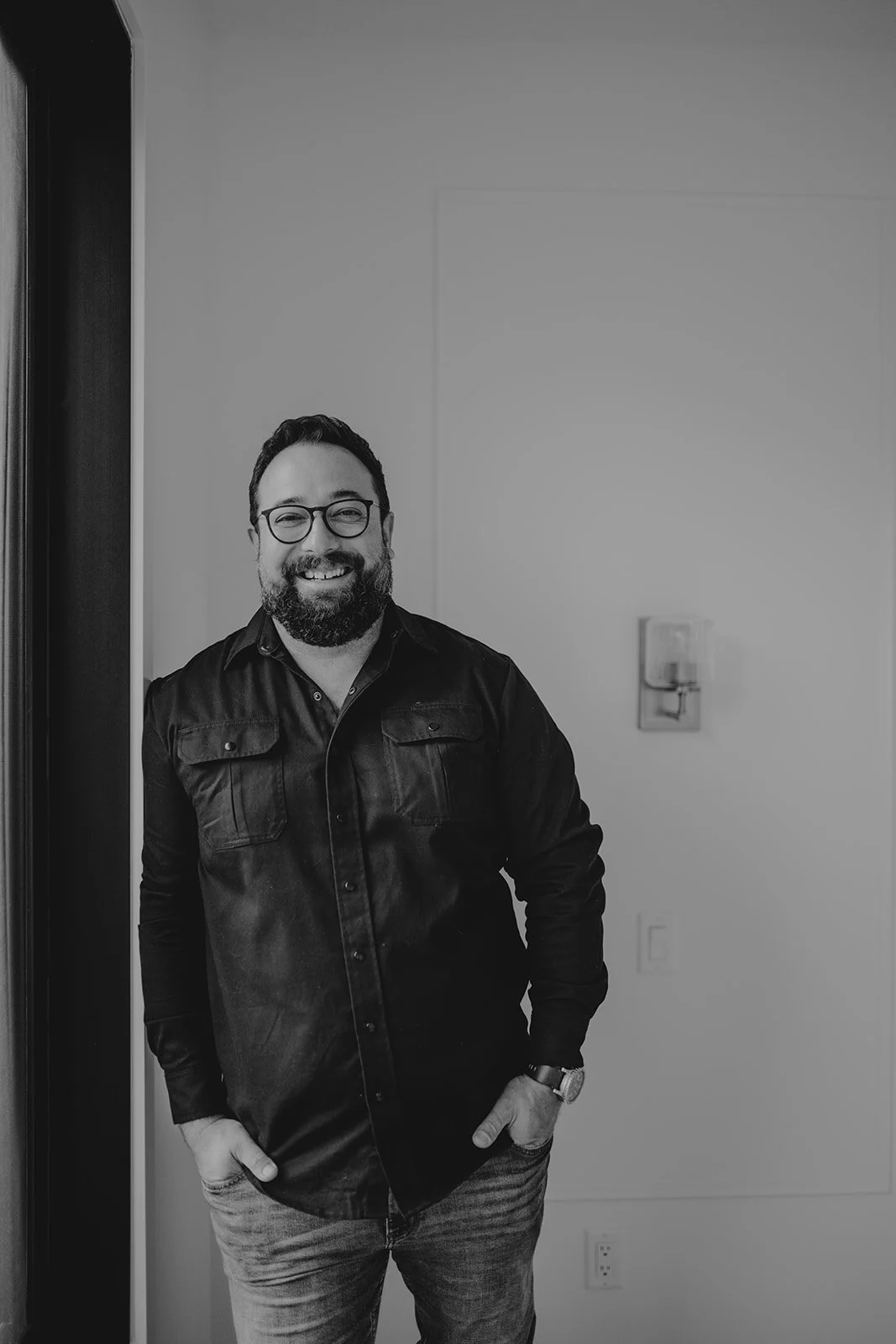 A smiling man with glasses and a beard, wearing a dark button-up shirt and jeans, standing indoors against a plain wall.