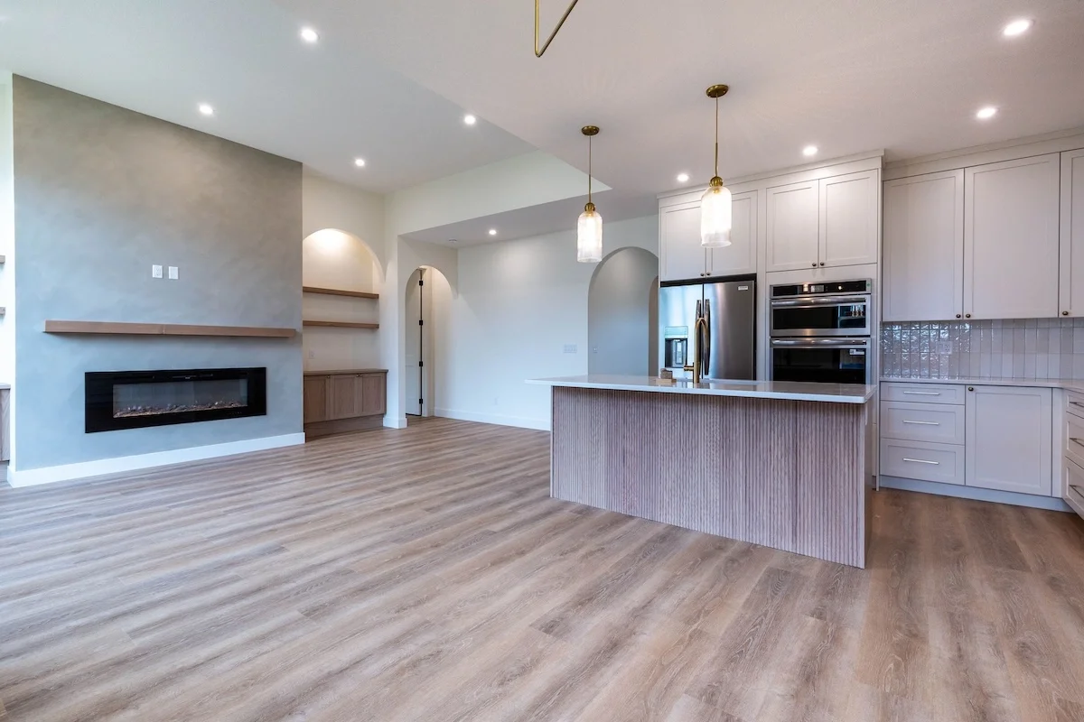 Modern open-concept living space with a fireplace, light wood flooring, and a kitchen with white cabinets, stainless steel appliances, and two pendant lights.