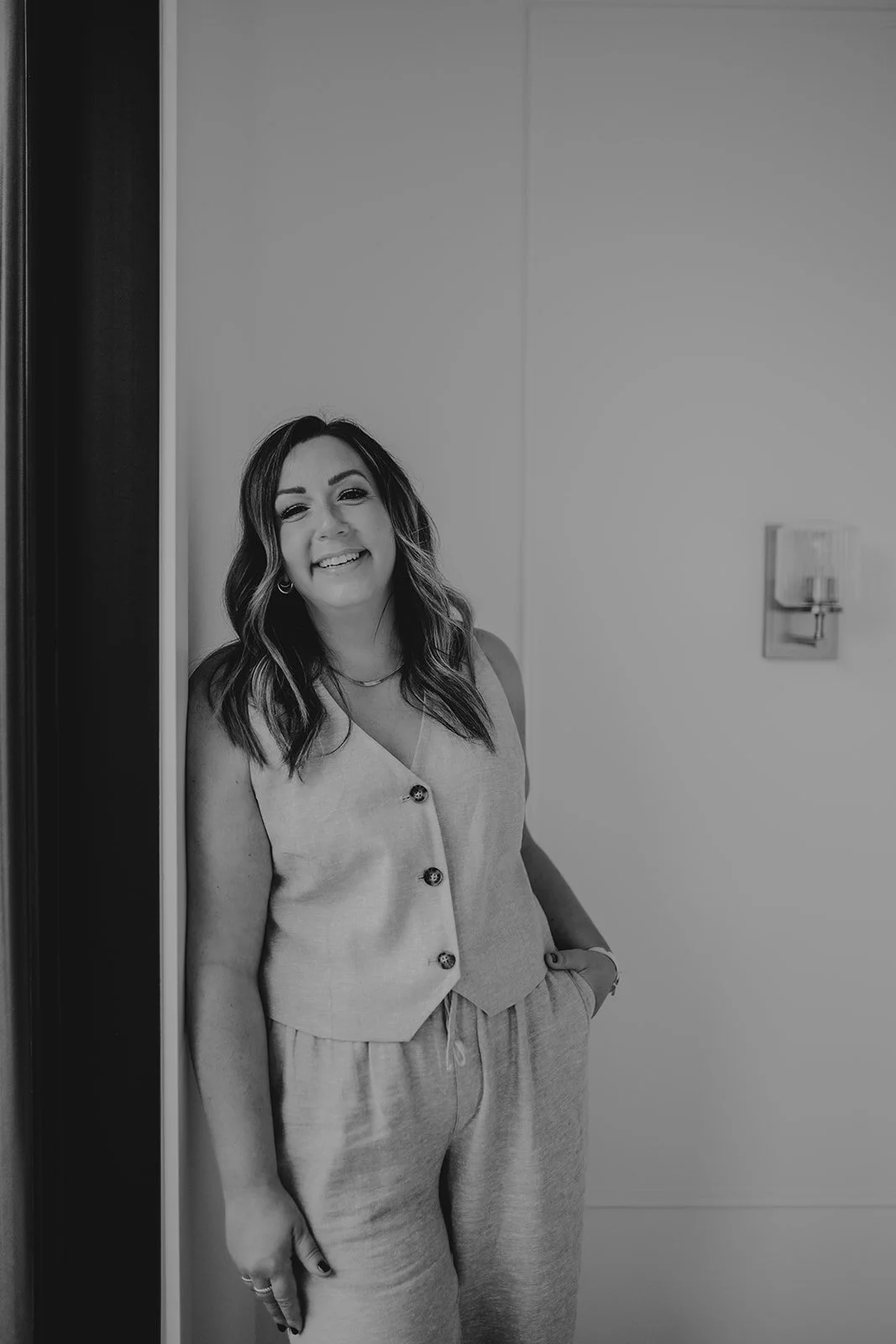 A woman standing and smiling in a doorway, wearing a sleeveless buttoned top and drawstring pants.