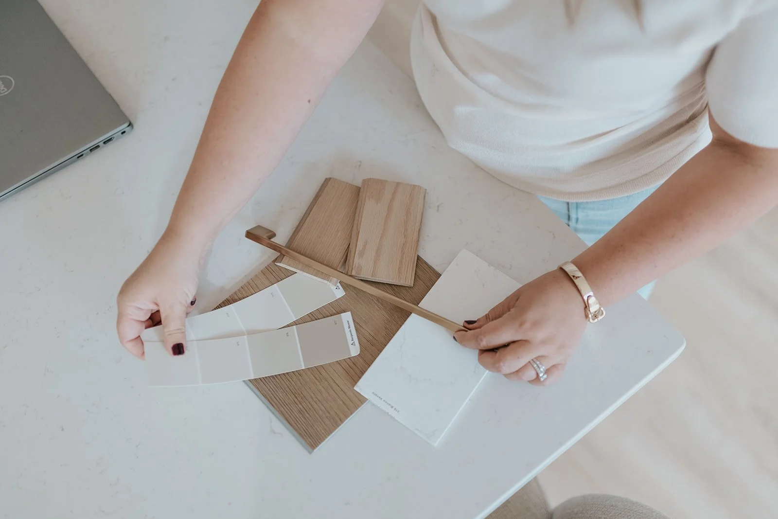 Person holding a color swatch card and a marble-patterned tile sample while standing at a white table with wood samples, a laptop, and other tile samples.