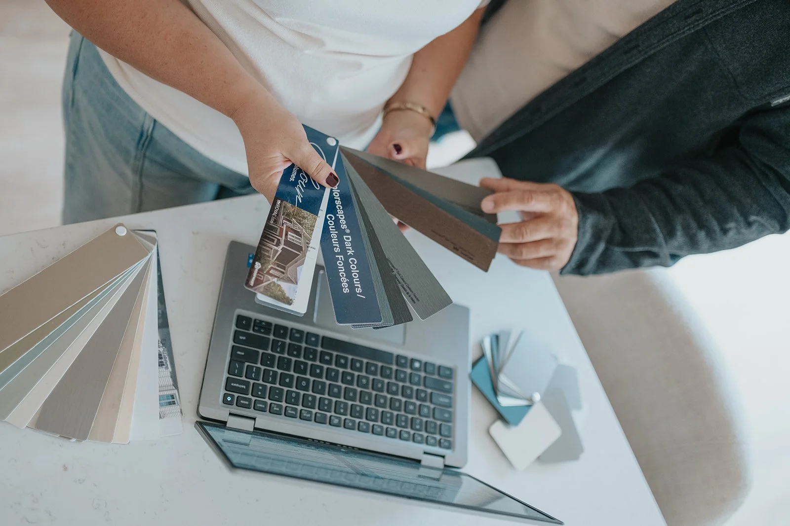 Two people comparing color samples and paint swatches at a white table with a laptop, sample cards, and color palettes.