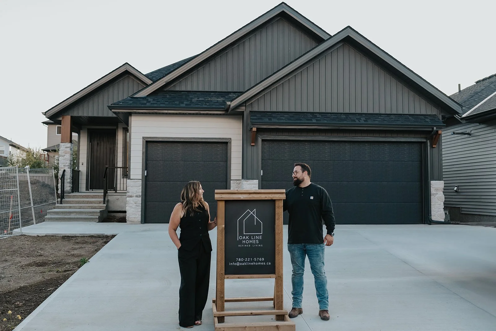 A man and woman standing outside a newly built house, next to a sign for Oak Line Homes. The house has a dark gray and white exterior with two garage doors, stairs, and a front porch.
