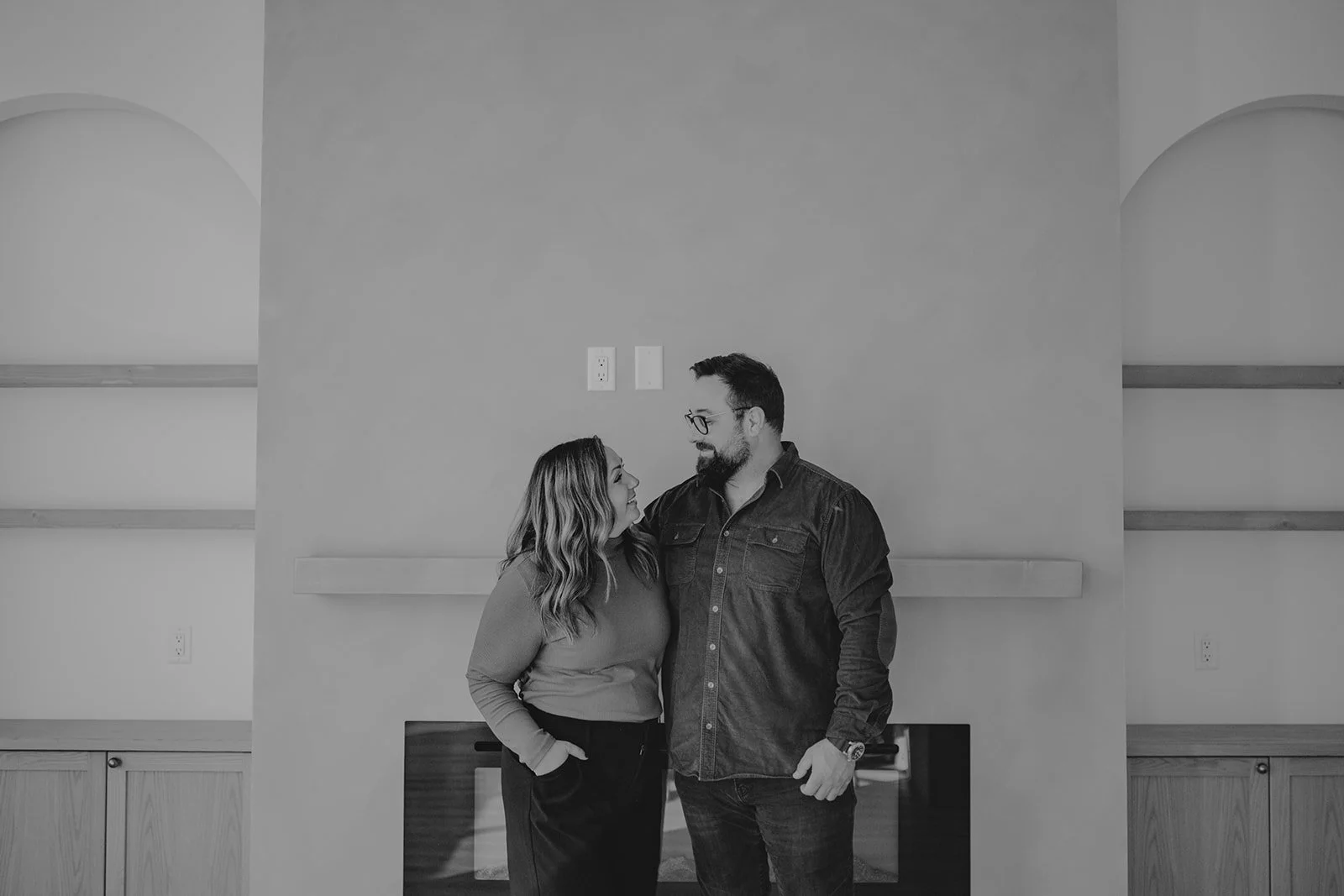 A Black and white photo of a man and woman looking at each other affectionately, standing in a room with minimal decor and a fireplace behind them.