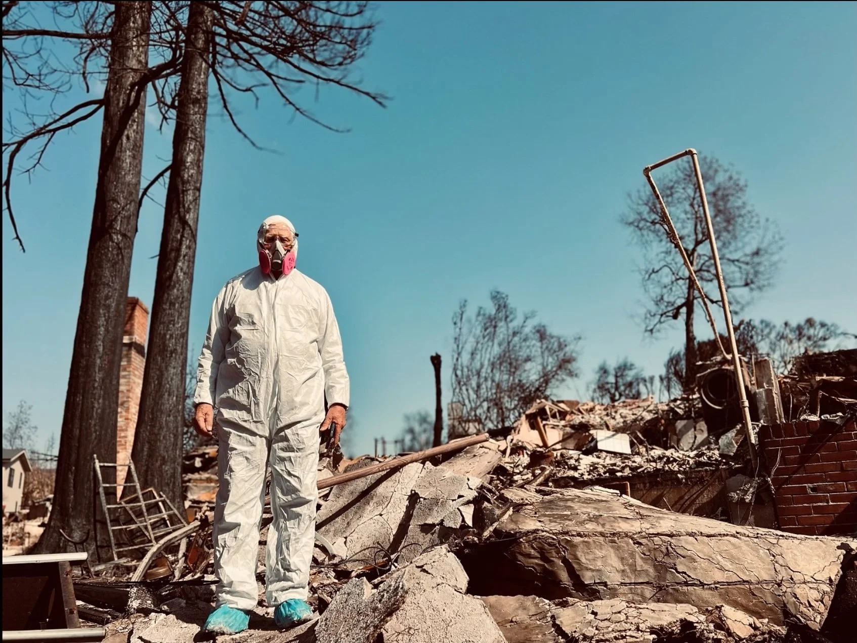 A man wearing a hazmat suit and ventilator, Gerhard Pichel, stands amid the ruins of his Pacific Palisades home destroyed by the January 7, 2025 Palisades Fire.