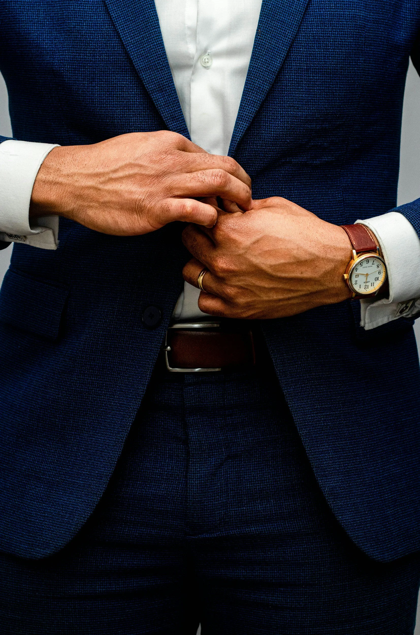 Close-up of a man adjusting his suit jacket. He is wearing a blue suit, white shirt, and a watch on his left wrist.