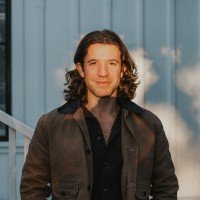 A young man with shoulder-length curly hair wearing a black shirt and a dark jacket, standing outdoors with a glass building and blue sky in the background.