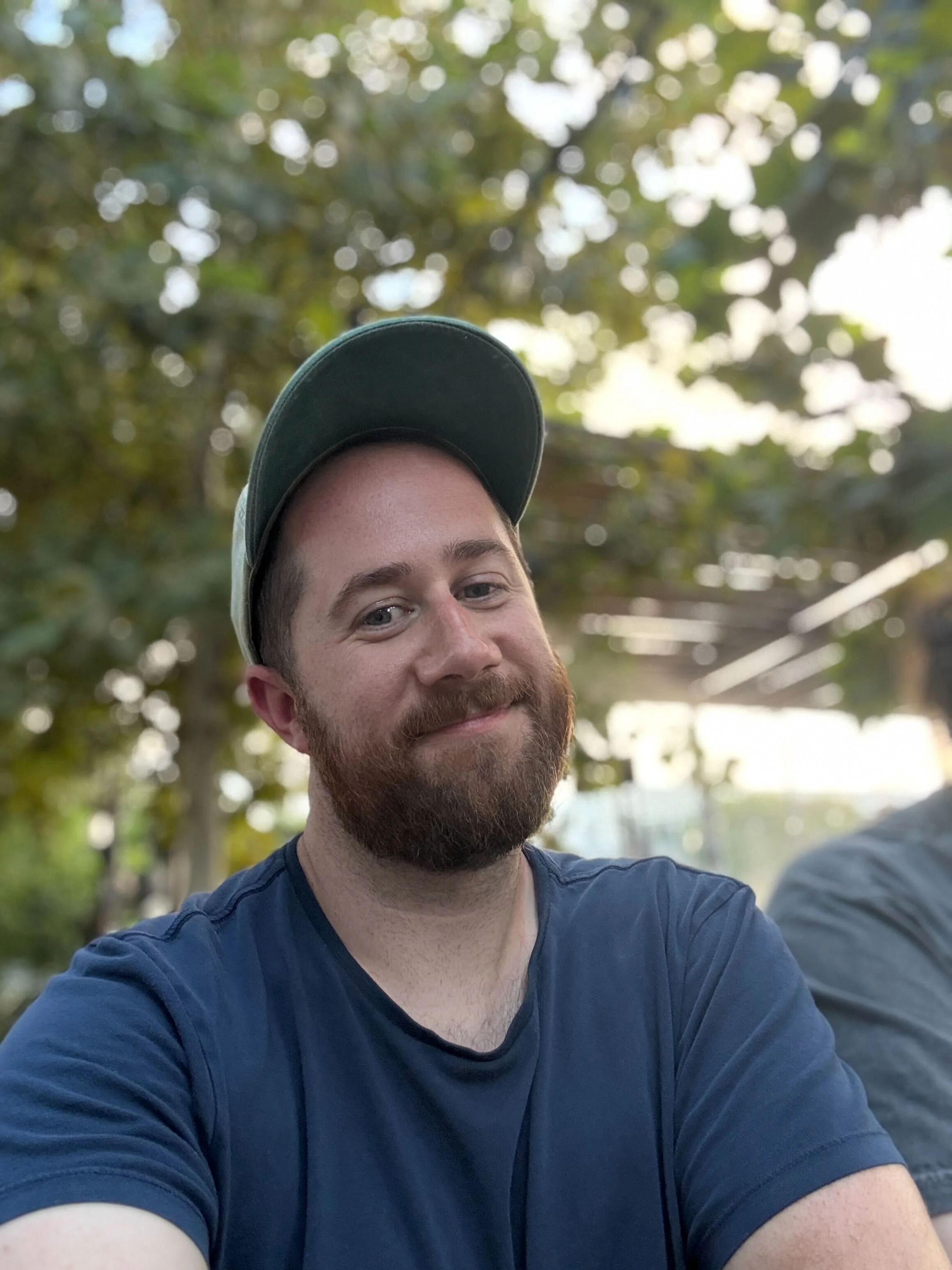 A man with a beard and mustache wearing a dark baseball cap and a blue t-shirt, smiling outdoors with blurred trees in the background.