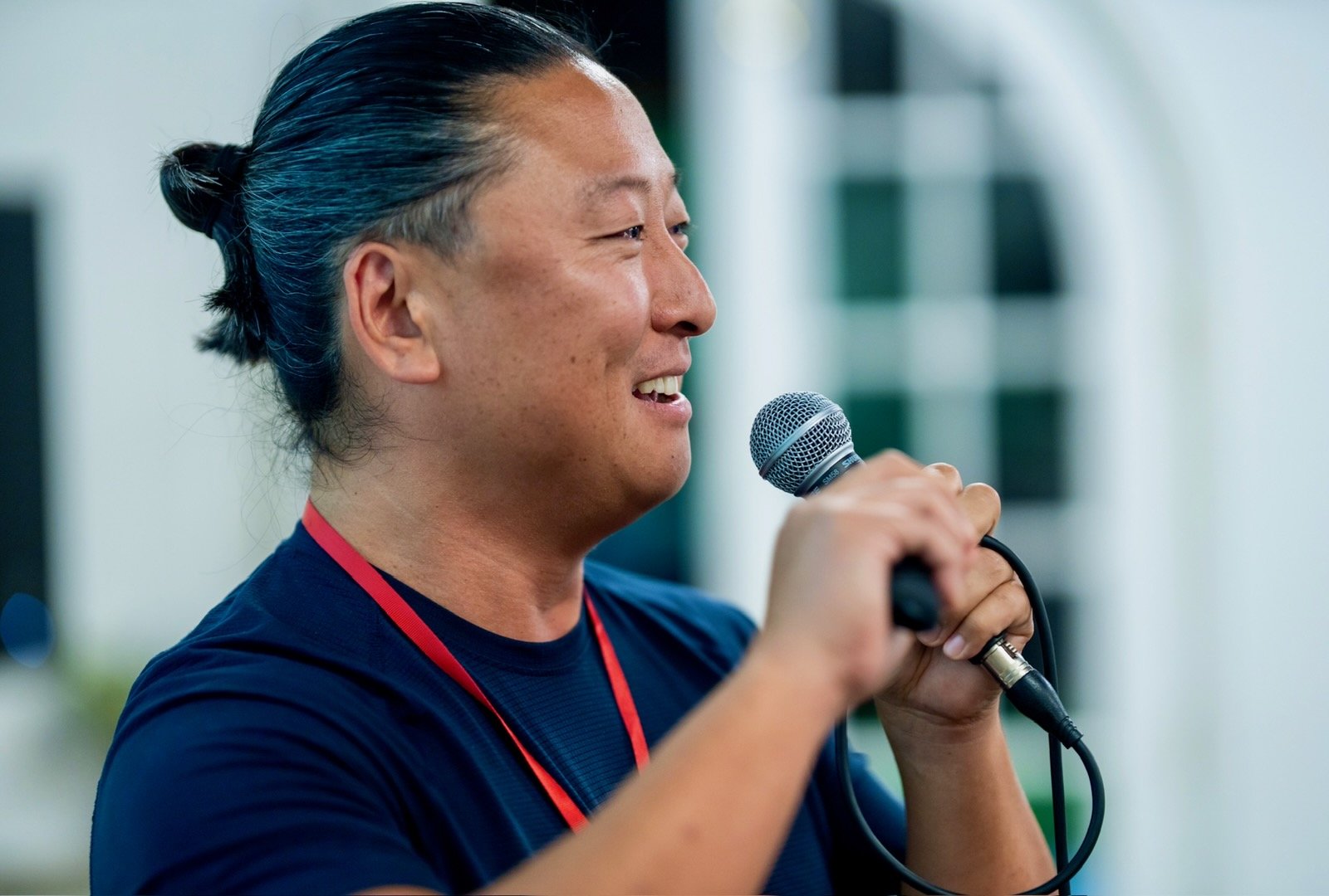 A person with dark hair tied back, holding a microphone and speaking or singing, smiling, wearing a dark shirt and a red lanyard, outdoors with a building in the background.