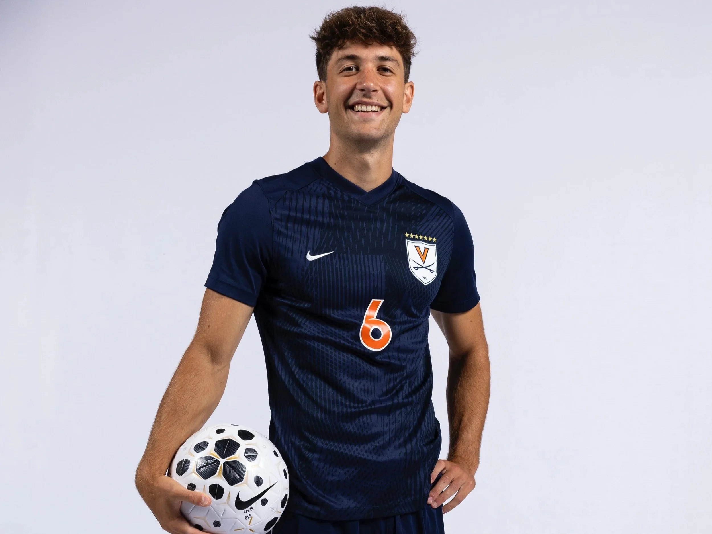 A young male soccer player wearing a navy blue jersey with the number 6, holding a soccer ball in his right hand, smiling against a plain white background.