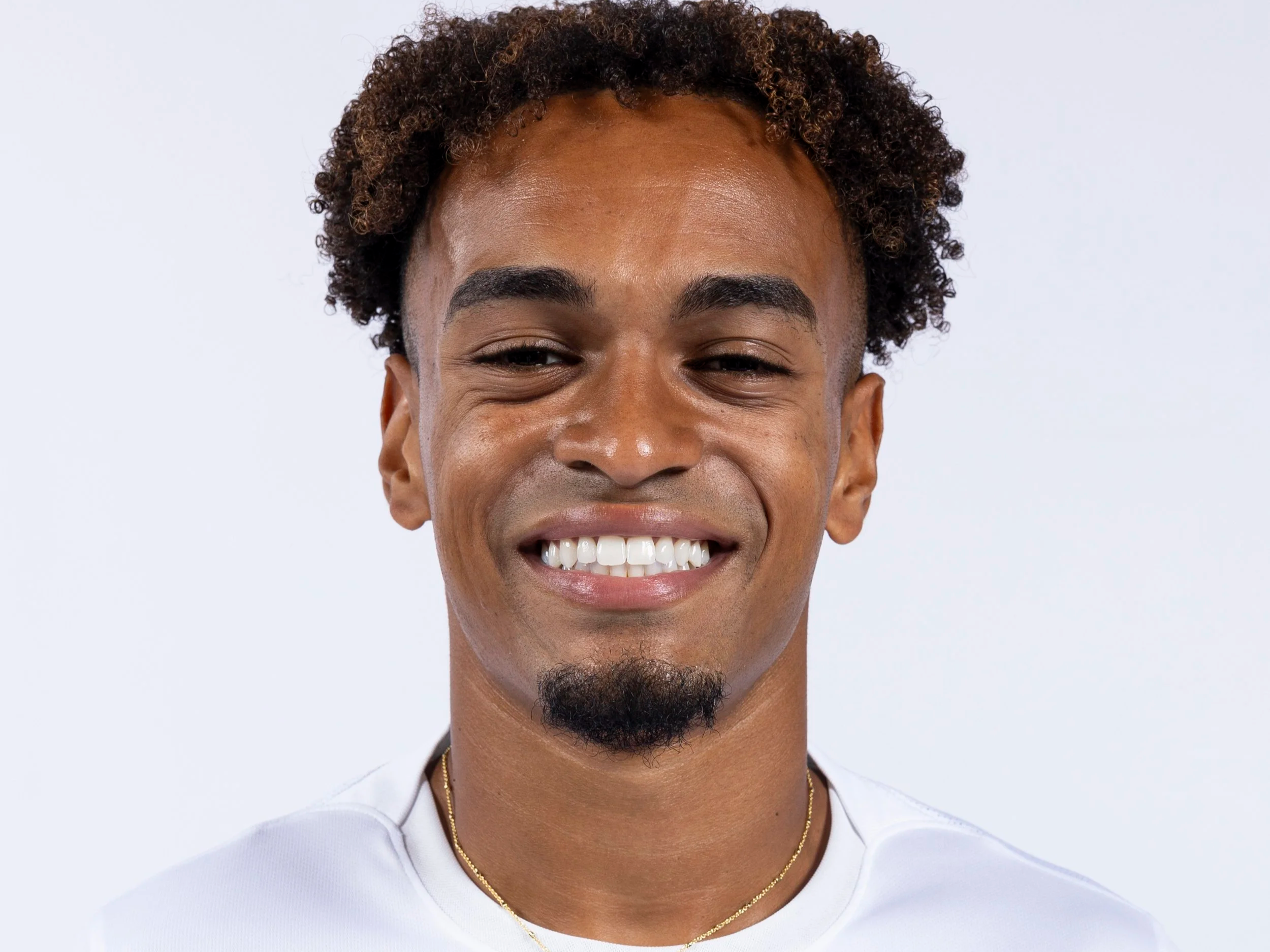Close-up portrait of a smiling young man with curly hair, brown skin, and facial hair, wearing a white shirt and a gold necklace.