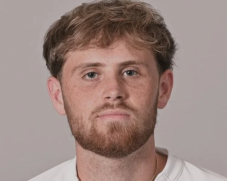 A young man with light brown hair, a beard, and blue eyes looking directly at the camera against a plain light gray background.