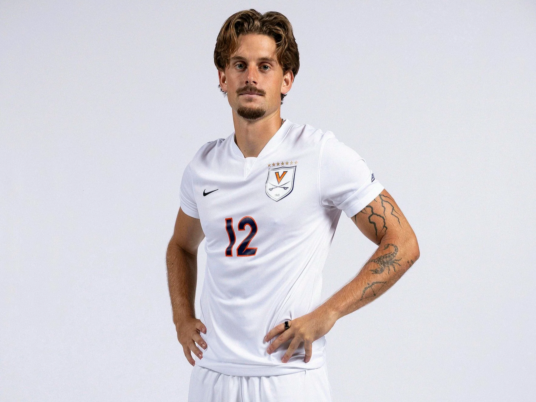 A male soccer player with light brown hair and a goatee, wearing a white jersey with the number 12 and a Virginia Cavaliers logo, standing with hands on hips against a plain white background.