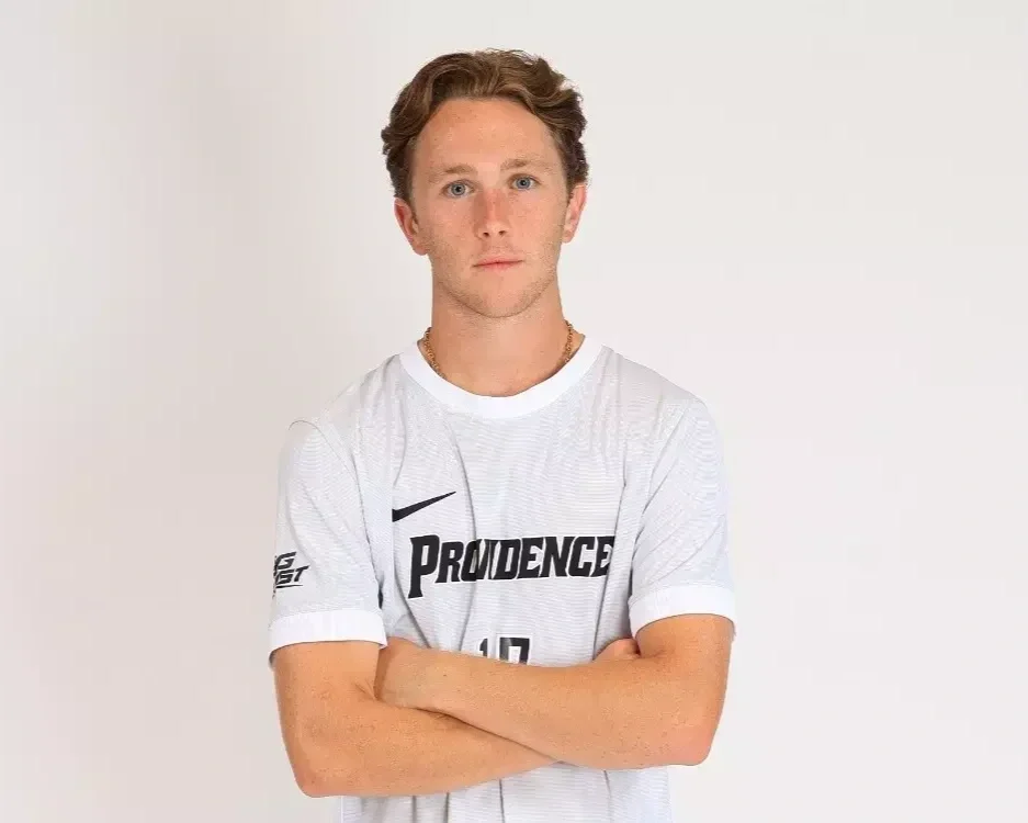 A young man with light brown hair and blue eyes crossing his arms, wearing a white Providence sports jersey against a plain white background.