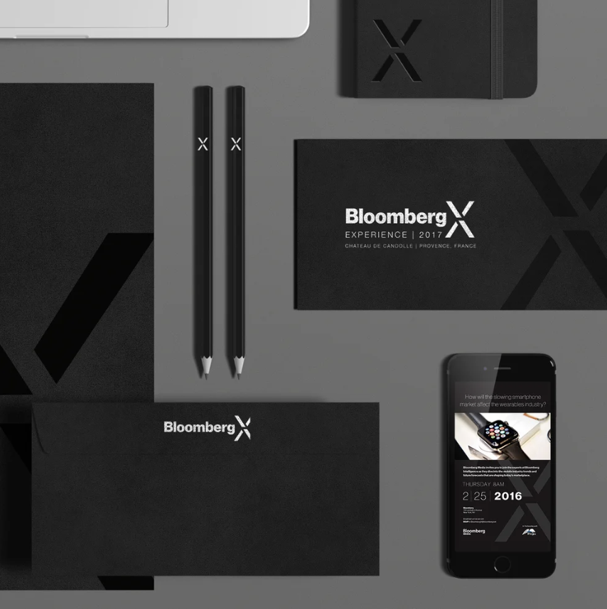 Flat lay of Bloomberg X-branded stationery and electronic devices, including two black pens, two black notebooks, a smartphone displaying a Bloomberg event, and a black carrying case, all on a gray background.