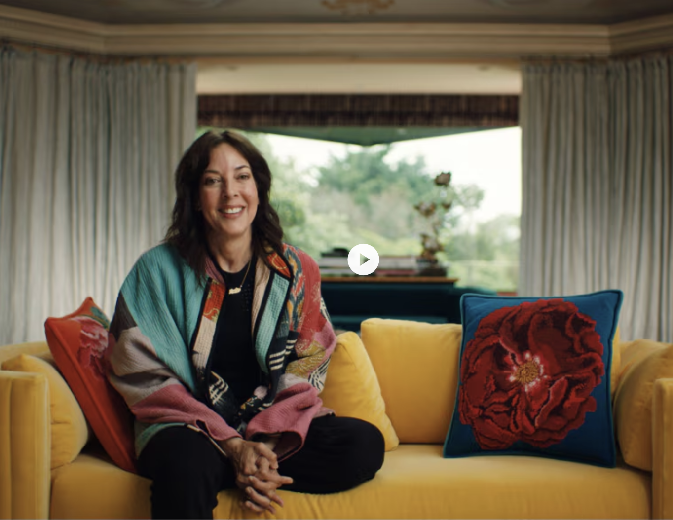 A woman (Leslie Blodgett) sitting on a yellow sofa with colorful pillows in a room with large windows and curtains, smiling at the camera from the Goldman Sachs Private Wealth Management testimonial series. 
