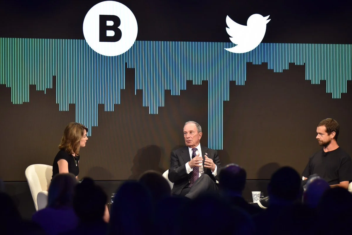 Panel discussion with three people (Mike Bloomberg, Emily Chang, Jack Dorsey) in front of a large screen displaying social media logos for Bloomberg and Twitter, and a wave pattern.
