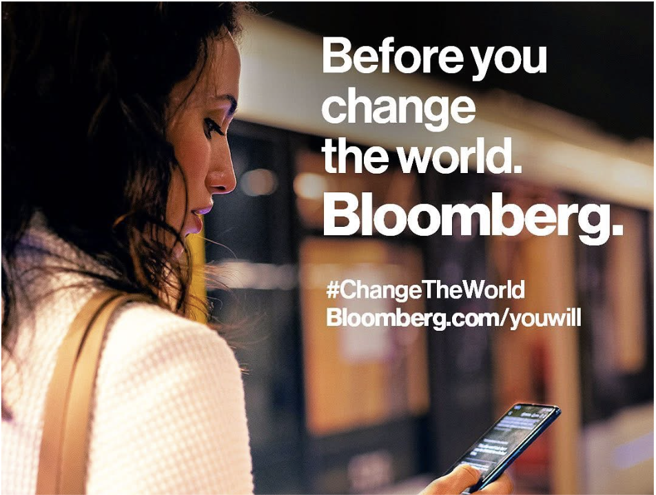 A woman with dark hair looking at her phone in a public space, with overlaid text that reads 'Before you change the world. Bloomberg.' from a Bloomberg Media global brand awareness campaign.
