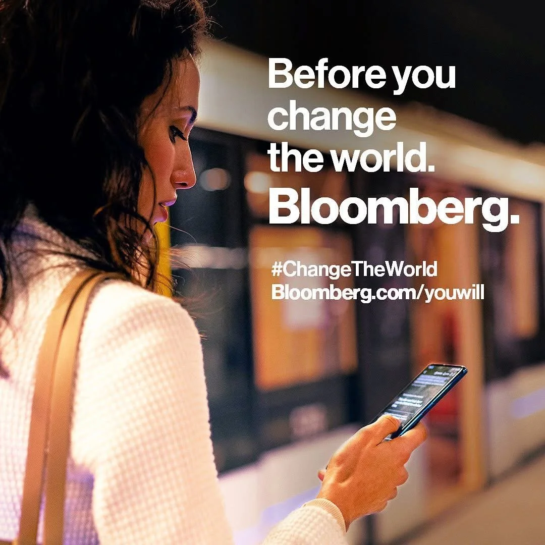 A woman with dark hair looking at her phone in a public space, with overlaid text that reads 'Before you change the world. Bloomberg.' from a Bloomberg Media global brand awareness campaign.
