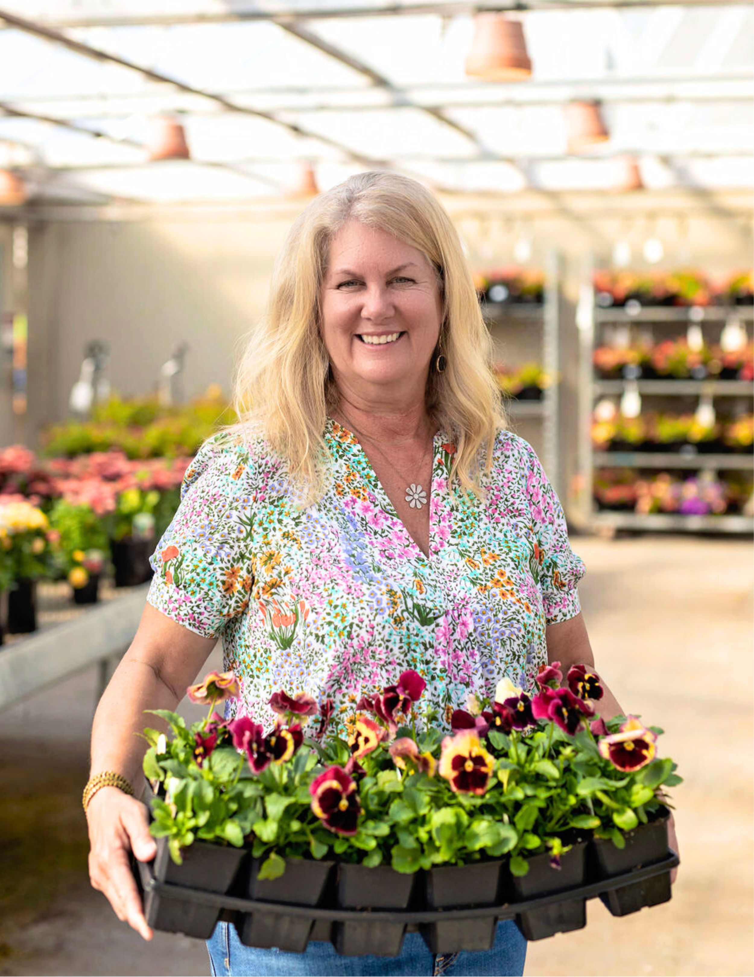 Cindy Funes holding a tray of pink, purple, and yellow pansies inside a garden center, with tables of potted plants in the background.