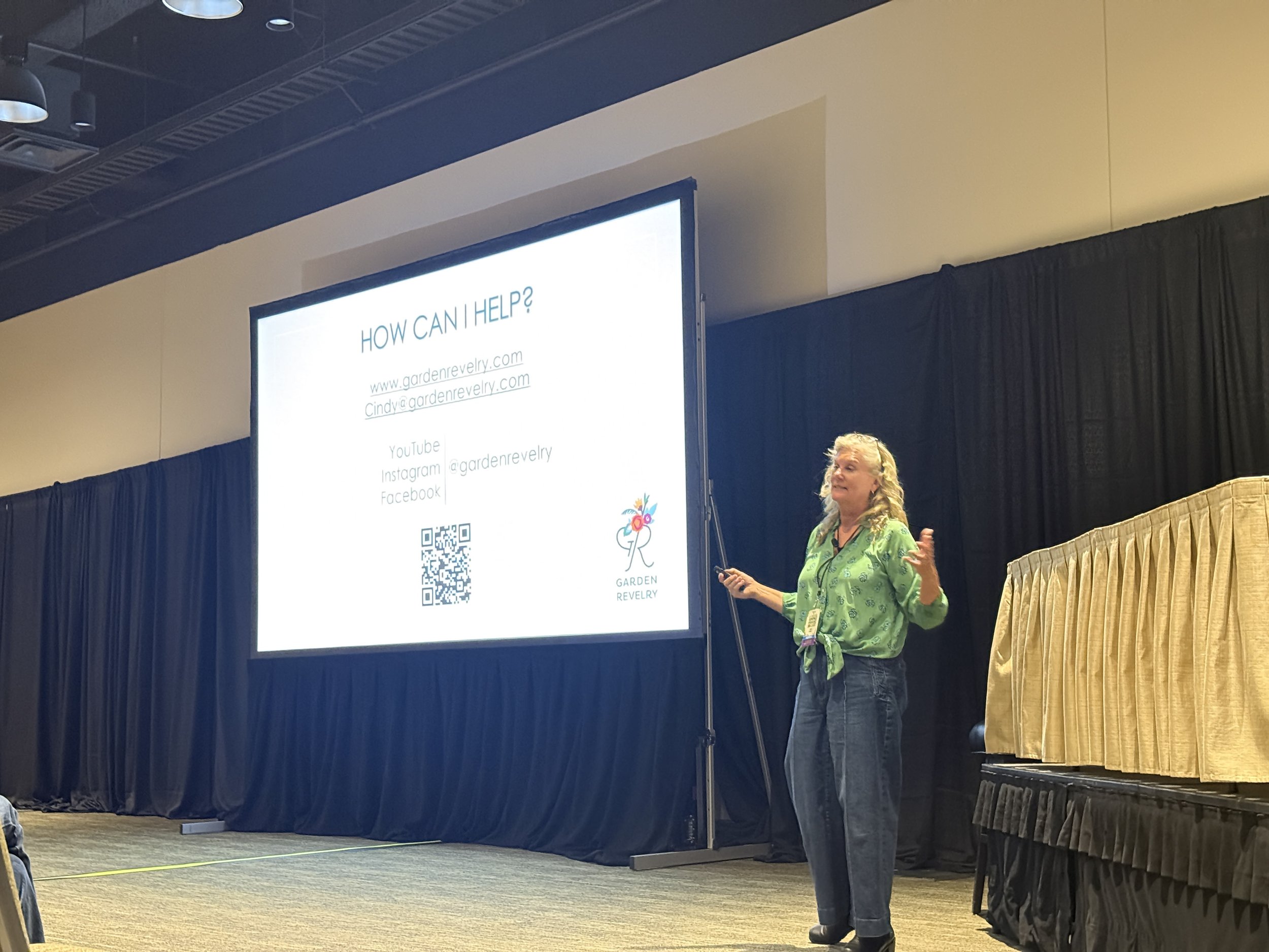 A woman with curly blonde hair is giving a presentation in front of a large screen that displays contact information and social media links for Garden Revelry. She is gesturing with her hands and wearing a green patterned shirt. The presentation slide includes a QR code and a flower logo.