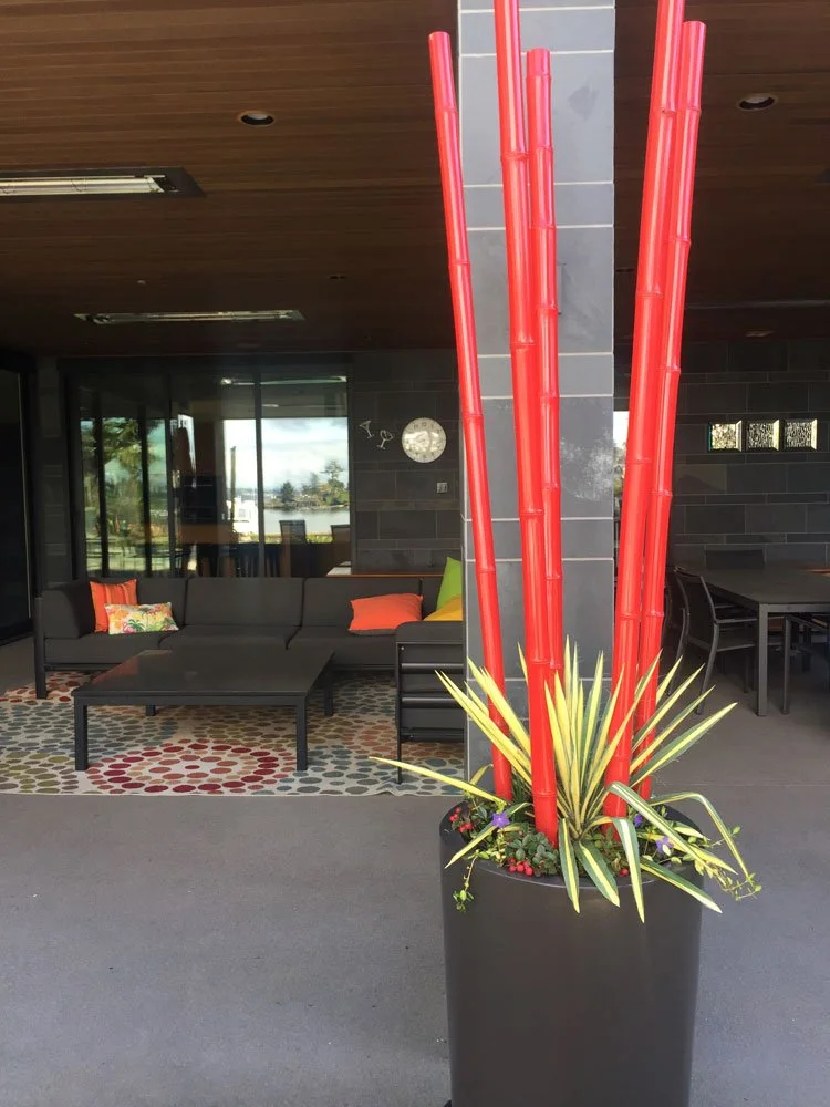 Indoor patio area with a black sectional sofa, colorful cushions, a patterned rug, and a tall black planter with red bamboo stalks and green plants.