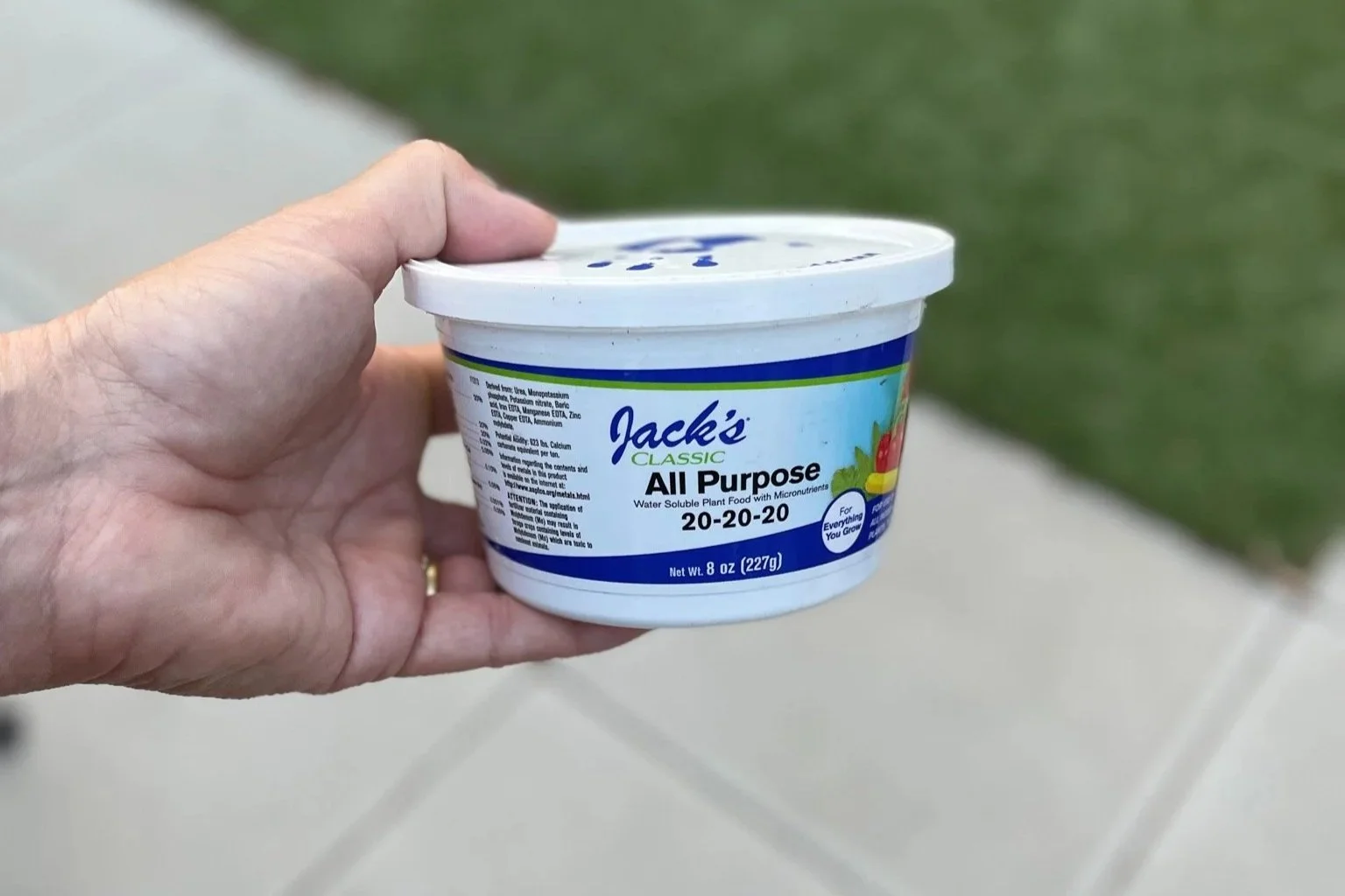 Cindy Funes' hand holding a tub of Jack's Classic All Purpose water-soluble plant food with micronutrients, 8 ounces, outdoors with a grassy area and sidewalk in the background.