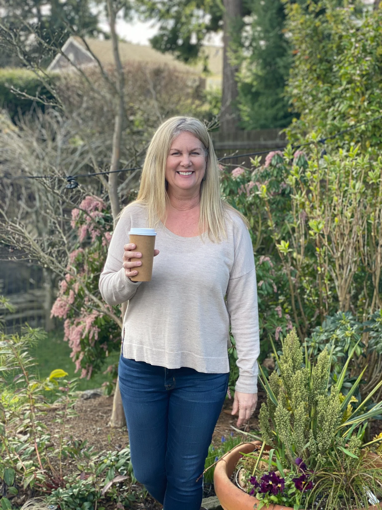 Woman with long blonde hair smiling, holding a coffee cup in a garden with green plants, pink flowers, and a large potted plant with purple flowers.