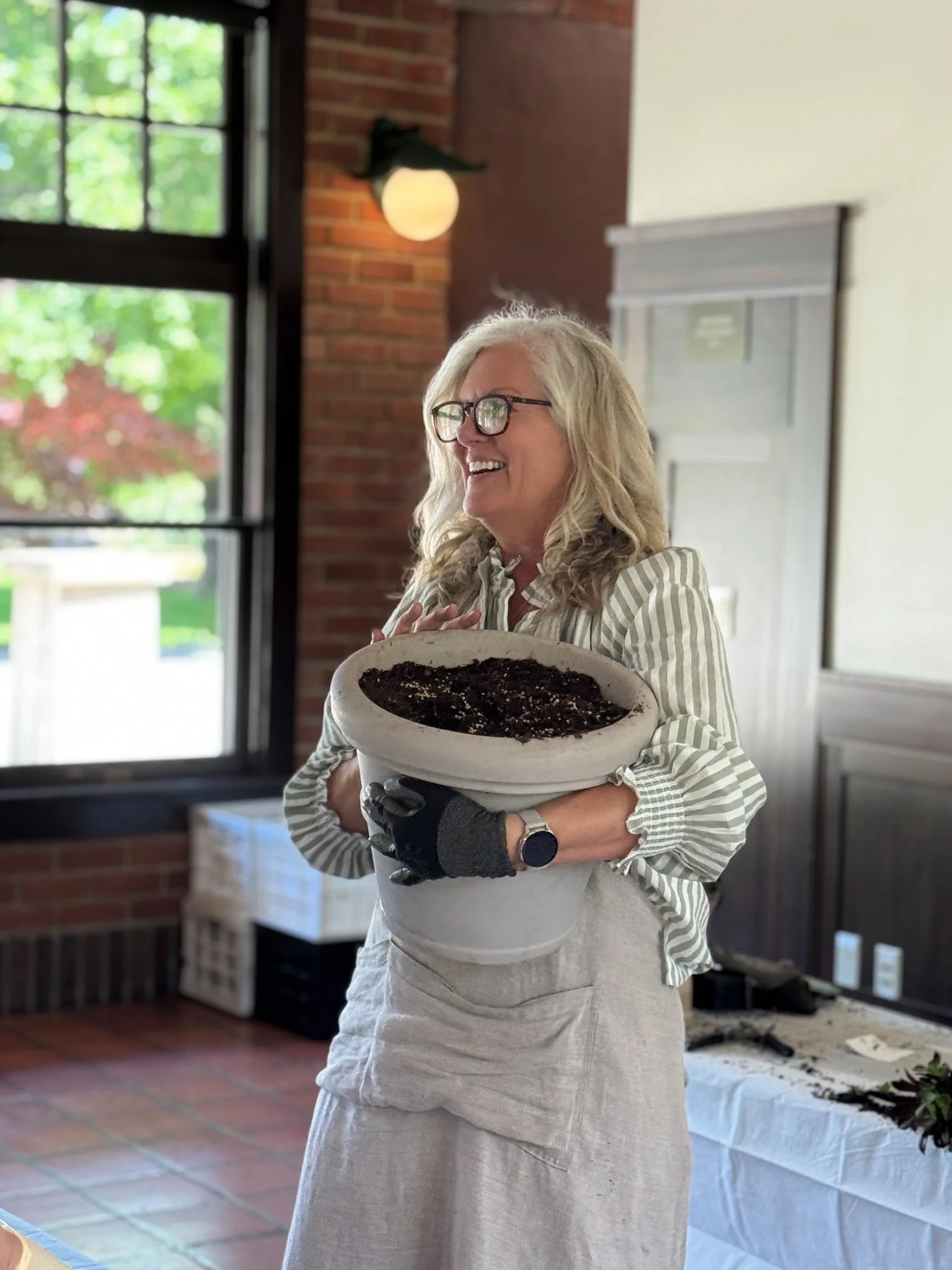 A woman with glasses and gray hair holding a bowl filled with dark soil, smiling indoors near a window and brick wall.