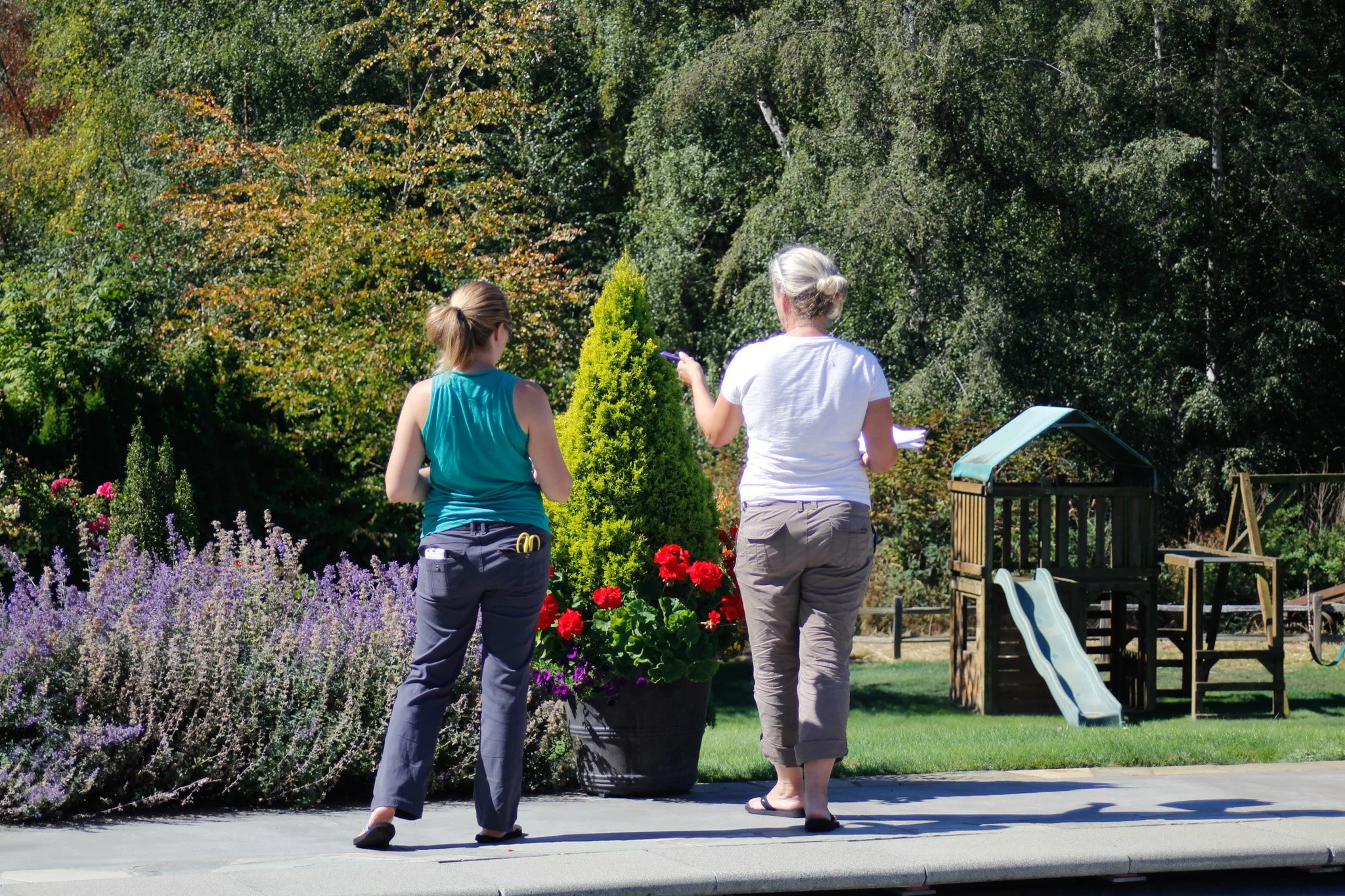 Two women are walking together in a garden, surrounded by colorful flowers and lush green trees, with a small slide in the background.