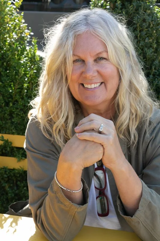 A smiling woman with long blonde curly hair wearing a gray jacket, sitting outdoors with sunlight on her face, green bushes in the background, and her hands clasped together.