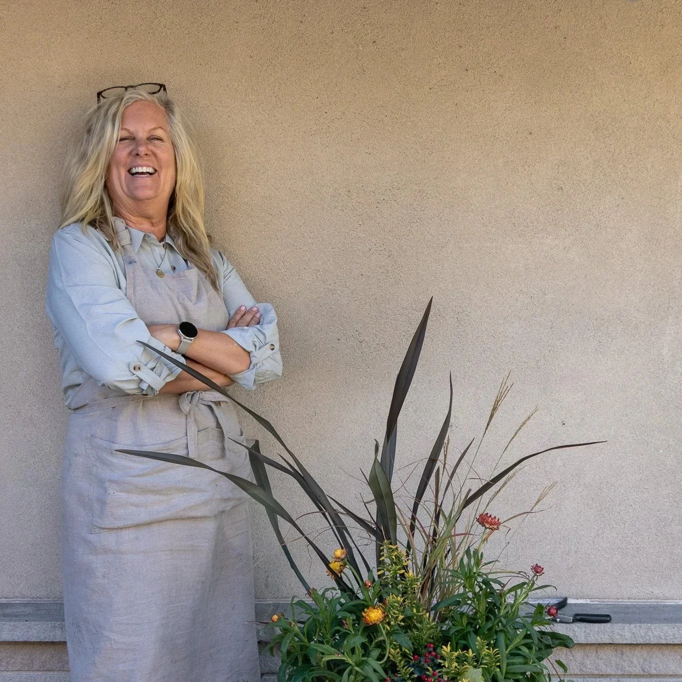 A woman with blonde hair smiling and standing with crossed arms next to a potted plant with long leaves and flowers, against a beige wall.