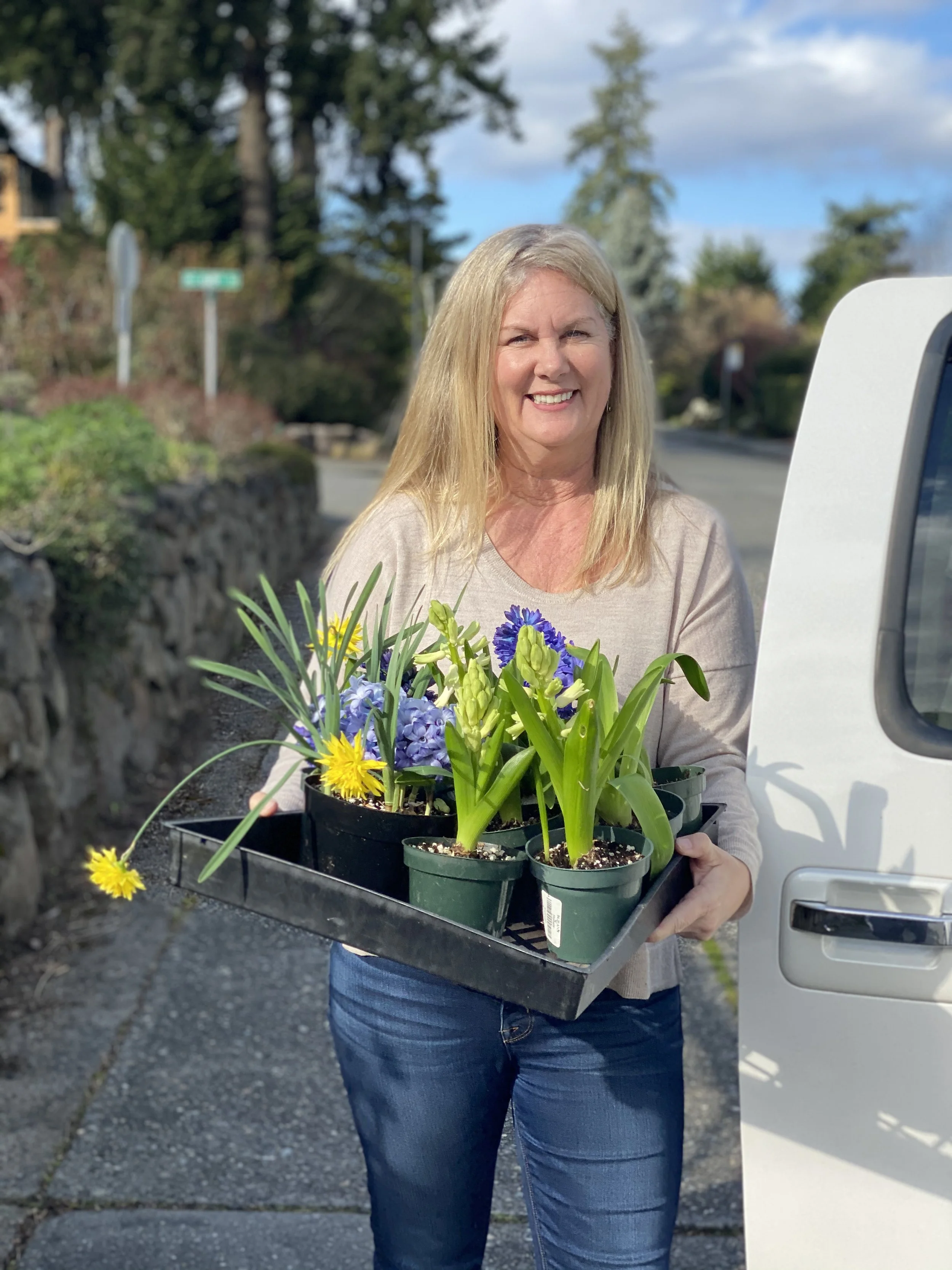 A woman smiling while holding a tray of potted flowers, including yellow and purple blooms, outdoors on a sunny day.