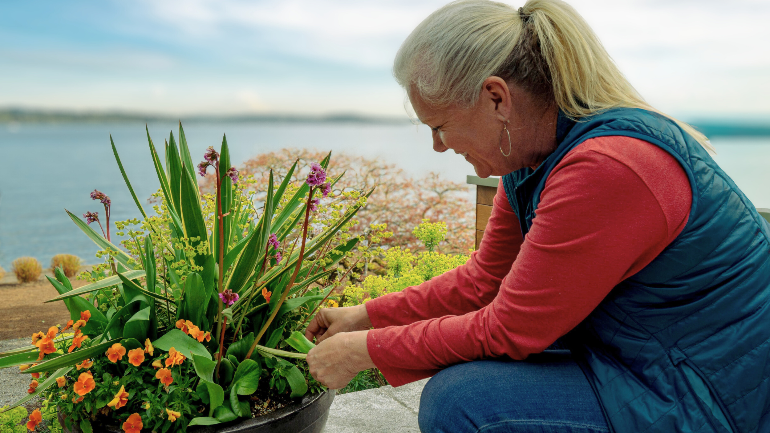 A woman with blonde hair, wearing a red long-sleeve shirt and blue vest, is tending to a potted garden with various colorful flowers near a body of water.