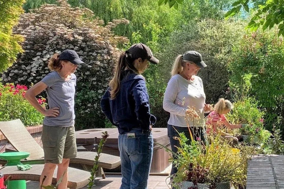Four women and a girl gardening in a lush backyard with trees and plants, some sitting on a patio table, one woman standing, the girl tending plants.