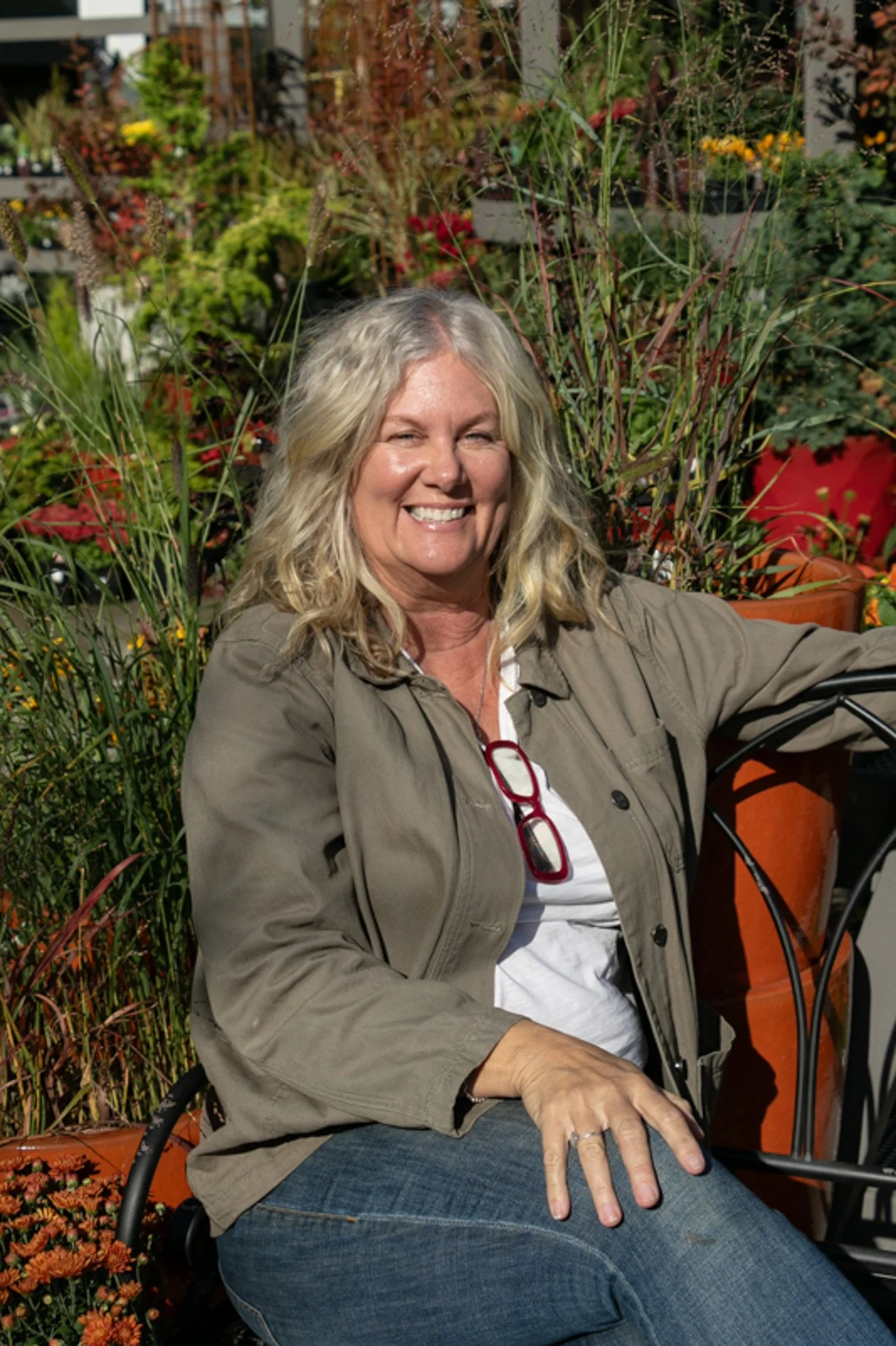 A smiling woman with blonde curly hair wearing a beige jacket and blue jeans sitting outdoors in a garden with colorful flowers.
