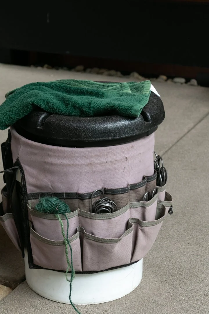 A black stool with a green cloth on top, surrounded by a pink knitting or crochet bag containing yarn and metal hooks, placed on a white round surface on a concrete floor.