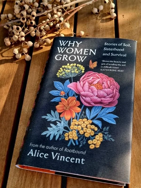 Close-up of a book titled "Why Women Grow: Stories of Soil, Sisterhood, and Survival" by Alice Vincent, with illustrated colorful flowers on the cover, placed on a wooden surface next to dried plant stems.