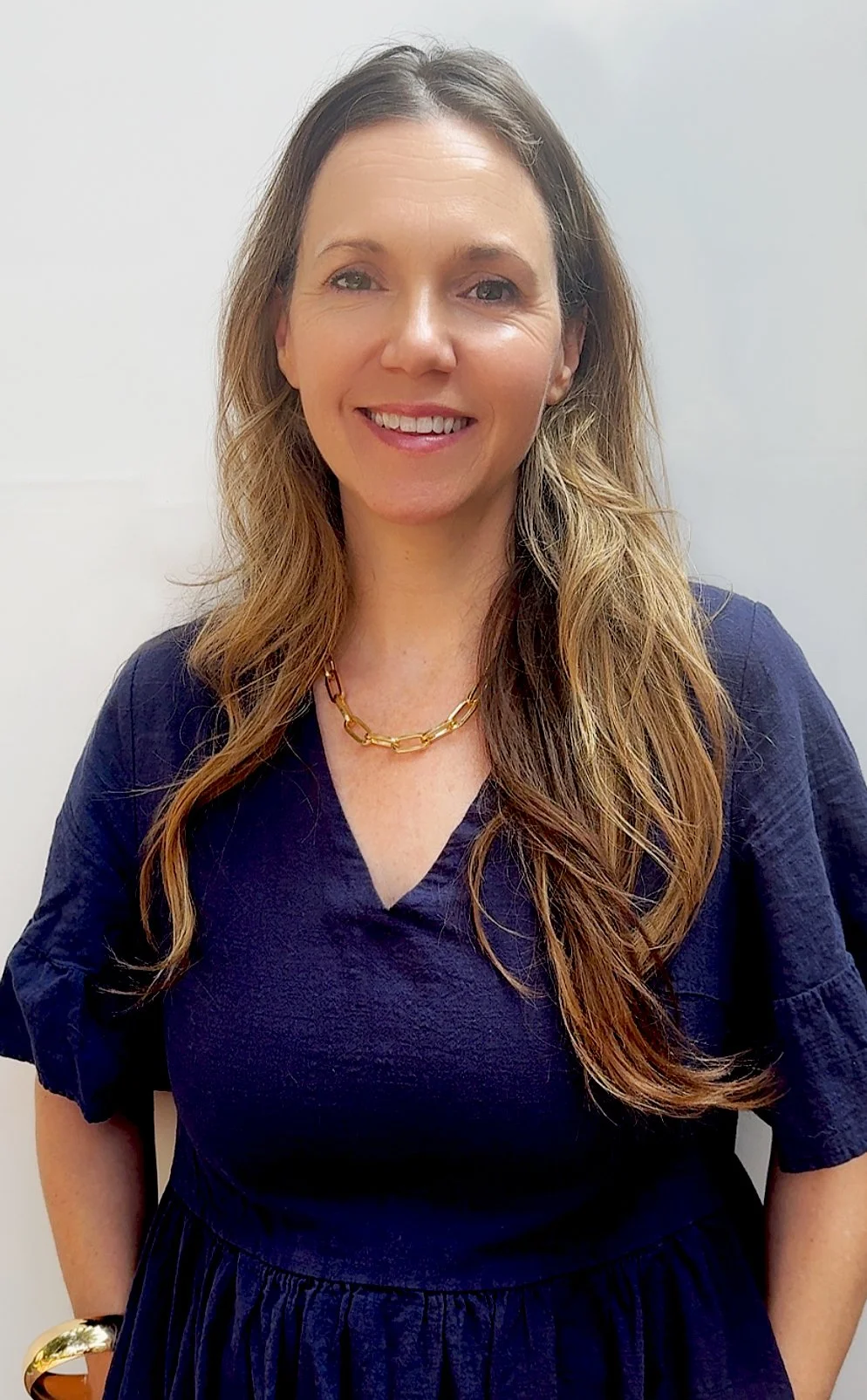 A woman with long wavy brown hair, wearing a navy blue dress, a gold chain necklace, a gold bracelet, and smiling in front of a plain white wall.
