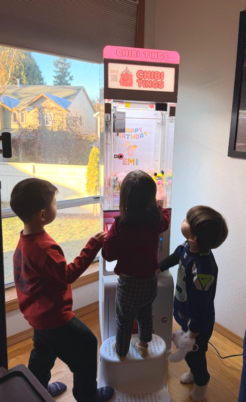 Three children playing a claw machine game indoors near a large window showing houses and trees outside. The machine is decorated with birthday messages and a pink sign reading 'Chibi Tings'.